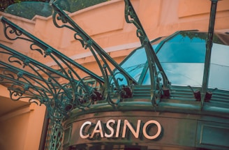 The image displays the entrance to a casino, characterized by detailed and ornate architectural elements with green metal structures and a sign that reads 'CASINO'. The background includes a glass dome and a peach-colored wall.