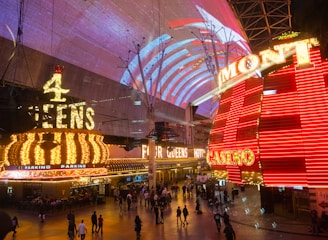 A bustling nighttime scene with illuminated casino signage towering over a busy pedestrian street. Neon lights in red and yellow highlight the names of various establishments. People are walking in different directions, enjoying the vibrant atmosphere.