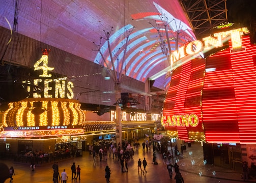 A bustling nighttime scene with illuminated casino signage towering over a busy pedestrian street. Neon lights in red and yellow highlight the names of various establishments. People are walking in different directions, enjoying the vibrant atmosphere.