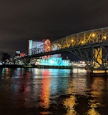 A brightly lit casino with neon signs and red lights is reflected in a river. A bridge stretches across the water, with an urban skyline visible in the background against an overcast night sky.