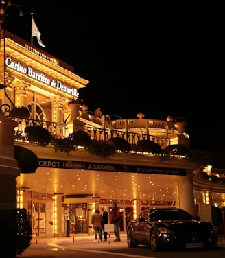 A luxurious casino at night, illuminated with warm, golden lights. The building features elegant architecture with columns and decorative elements. A few people are walking towards the entrance, and there are a couple of parked cars nearby.