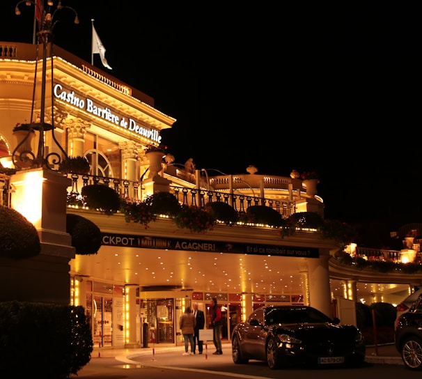 A luxurious casino at night, illuminated with warm, golden lights. The building features elegant architecture with columns and decorative elements. A few people are walking towards the entrance, and there are a couple of parked cars nearby.
