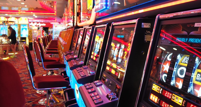 A brightly lit casino interior featuring a row of colorful slot machines. The neon lights create a vibrant atmosphere, with patterned carpeting adding to the decor. Stools are aligned neatly in front of each machine.