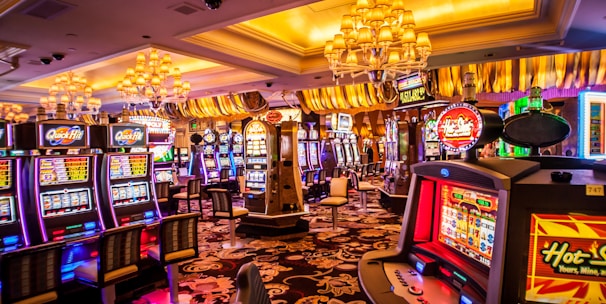 A vibrant casino interior filled with rows of colorful slot machines, each illuminated with bright lights and displays. The ceiling is adorned with elegant chandeliers and golden drapes, creating a luxurious atmosphere. The patterned carpet adds to the elaborate design of the space.