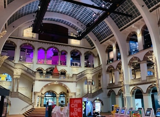 The interior of a large, ornately designed building with arched ceilings and intricate architectural details. The space features gaming machines lined up along the floor, under a high ceiling with a grid pattern. Lighting in purple and white tones enhances the ambiance.