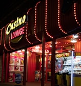 A brightly lit casino lounge entrance adorned with neon lights. The exterior features colorful signage including the words 'Casino' and 'Lounge' in vivid colors, with additional lights framing the building. Inside, a glimpse of a claw machine filled with plush toys and arcade games is visible. The area exudes an inviting yet bustling atmosphere typical of entertainment venues.