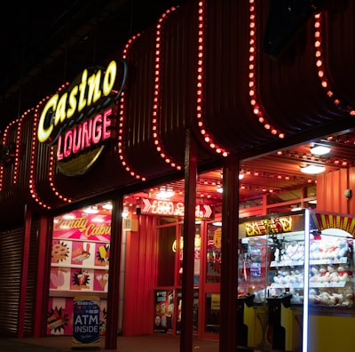 A brightly lit casino lounge entrance adorned with neon lights. The exterior features colorful signage including the words 'Casino' and 'Lounge' in vivid colors, with additional lights framing the building. Inside, a glimpse of a claw machine filled with plush toys and arcade games is visible. The area exudes an inviting yet bustling atmosphere typical of entertainment venues.