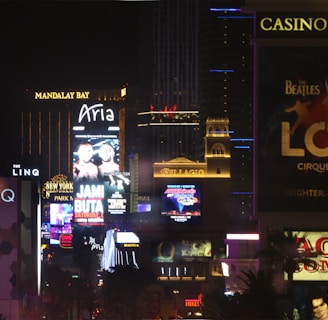 Bright city lights and large illuminated signs dominate a bustling urban skyline. Numerous advertisements and marquees for famous hotels and casinos are visible, showcasing entertainment and attractions in a vibrant nightlife setting.