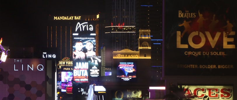 Bright city lights and large illuminated signs dominate a bustling urban skyline. Numerous advertisements and marquees for famous hotels and casinos are visible, showcasing entertainment and attractions in a vibrant nightlife setting.