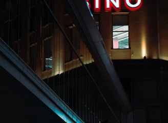A night scene featuring a building with illuminated neon signage displaying 'CASINO'. The environment is urban with reflections on glass windows and glowing street traffic lights below. The atmosphere is dimly lit, with artificial lighting casting vibrant colors.