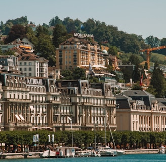 An elegant lakeside town with grand architecture featuring historic hotels and a casino. The buildings are set against a backdrop of lush green hills, and the foreground includes a marina with several yachts docked. The scene is characterized by vibrant greenery and a serene waterfront atmosphere.