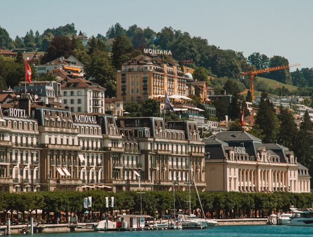 An elegant lakeside town with grand architecture featuring historic hotels and a casino. The buildings are set against a backdrop of lush green hills, and the foreground includes a marina with several yachts docked. The scene is characterized by vibrant greenery and a serene waterfront atmosphere.