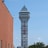 A tall tower with a modern design features the large vertical red letters spelling 'CASINO'. The structure has a sleek, metallic appearance with various horizontal and vertical lines. The sky is mostly clear with a few clouds. In the foreground, part of a brick building is visible, along with some trees and buildings in the distance.