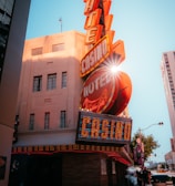 A vibrant city scene featuring a tall, illuminated casino sign with retro-style neon lights. The sign dominates the building facade with the words 'CASINO' and 'HOTEL'. Sunlight peeks through, creating a dramatic lens flare effect. The street is lively, with pedestrians casually walking and a hint of urban hustle.