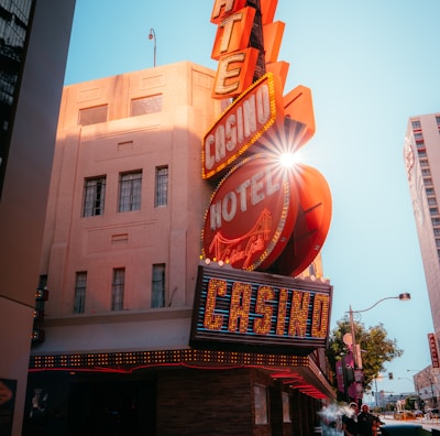 A vibrant city scene featuring a tall, illuminated casino sign with retro-style neon lights. The sign dominates the building facade with the words 'CASINO' and 'HOTEL'. Sunlight peeks through, creating a dramatic lens flare effect. The street is lively, with pedestrians casually walking and a hint of urban hustle.